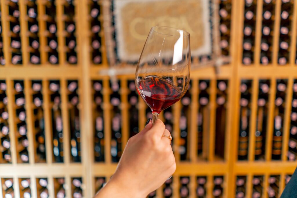 Calice di vino rosso in primo piano con cantina sullo sfondo durante una degustazione.